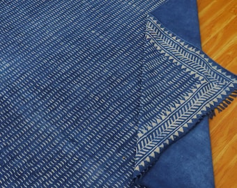 Op de afbeelding: Een blauwe katoenen stof met een wit geometrisch patroon. De stof heeft een gefranst randje en is over zichzelf gevouwen.