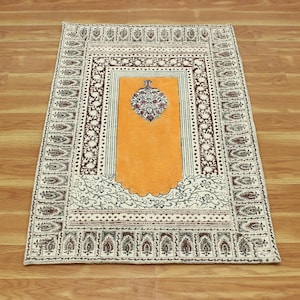Puede incluir: Una alfombra de oración rectangular con un centro naranja y un borde decorativo. El borde presenta un patrón repetitivo de diseños florales y geométricos en tonos marrón, blanco y crema. La alfombra está colocada sobre un suelo de madera.