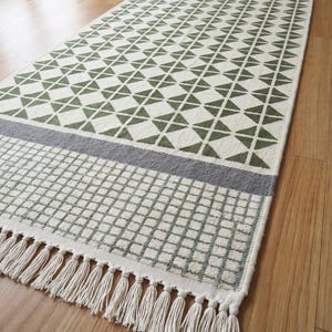 May include: A long rectangular runner rug featuring a repeating olive green geometric triangle pattern on a cream background. The rug includes a grey horizontal band, a grid-patterned section, and cream-colored fringe tassels at the ends.