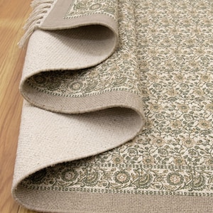 Puede incluir: Alfombra beige enrollada con un estampado floral en tonos verdes y beige. La alfombra tiene un borde beige y flecos en el borde. La alfombra está colocada sobre un suelo de madera.