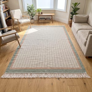 Puede incluir: Una alfombra rectangular con un centro de color crema y una cuadrícula de líneas negras finas. La alfombra está bordeada por una banda ancha de color rosa apagado seguida de una banda más ancha de color verde salvia, con flecos largos.