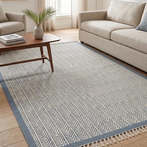 Puede incluir: Una alfombra rectangular con borde azul y un patrón de espiga repetido en blanco y gris. La alfombra tiene flecos en los extremos cortos y está colocada sobre un suelo de madera.