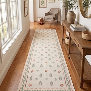 Pode incluir: Um tapete passadeira retangular longo com uma base de cor creme e um padrão repetitivo de folhas verde-sálvia e pequenos grupos de flores de cor terracota. O tapete é finalizado com uma borda decorativa e franjas longas em ambas as pontas.