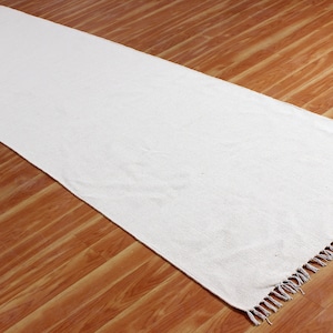Puede incluir: Una alfombra larga y rectangular de color blanco roto con un borde con flecos. La alfombra está hecha de un material tejido y se muestra sobre un suelo de madera. El color neutro y el diseño sencillo de la alfombra la hacen adecuada para varios estilos de interior.