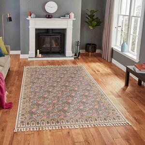 Può includere: Un tappeto rettangolare caratterizzato da un fitto motivo floreale e botanico nei toni del rosa, blu e verde. Il tappeto presenta un bordo decorativo con un motivo a foglie ripetuto e frange bianche sui lati corti, posato su un pavimento.