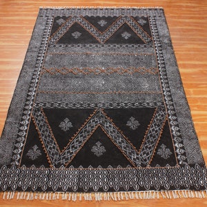 Puede incluir: Alfombra de patrones negros y blancos con acentos marrones. La alfombra tiene un diseño geométrico con flecos en los bordes.