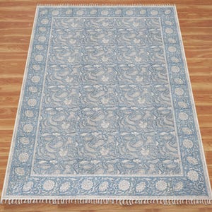 Könnte beinhalten: Ein rechteckiger Teppich mit einem hellblauen und weißen Blumen- und Paisley-Muster. Der Teppich hat eine breite dekorative Bordüre mit sich wiederholenden Blumenmotiven und ein zentrales Feld mit komplexen botanischen Designs, abgeschlossen mit weißen Fransen.