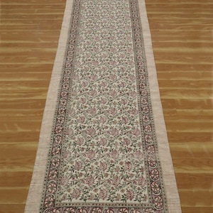 May include: A white cotton runner with a floral print in shades of pink and green. The runner has a border with a repeating pattern and fringe on the ends.