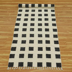 Puede incluir: Un tapete de corredor a cuadros blanco y negro con flecos en los extremos. El tapete está hecho de un material natural y tiene un diseño geométrico simple.