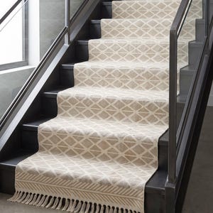 Puede incluir: Una alfombra larga para escaleras con un patrón de diamantes repetitivo en color beige y blanco roto. La alfombra está instalada sobre escalones de color gris oscuro e incluye un borde decorativo con rayas diagonales y flecos en la base.