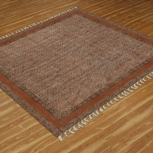 Puede incluir: Una alfombra de color marrón y negro con un borde de color óxido y flecos blancos. La alfombra está colocada sobre un suelo de madera.