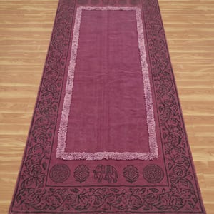 Puede incluir: Una alfombra rectangular de color magenta con un panel central bordeado por un fleco texturizado. El marco exterior muestra un patrón oscuro impreso de enredaderas florales, medallones circulares y un motivo de elefante, con borlas blancas.