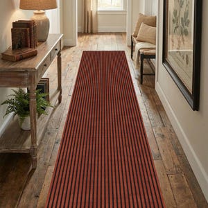 May include: A long, rectangular runner rug featuring a repeating pattern of vertical black stripes against a burnt orange background, finished with short, white fringe tassels along the narrow ends of the textile.