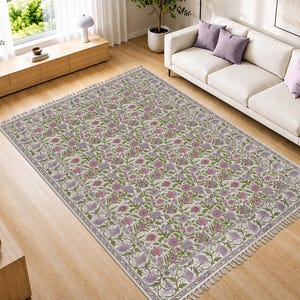 Op de afbeelding: Een rechthoekig vloerkleed met een crèmekleurige basis en een ingewikkeld bloemenpatroon in tinten roze, lavendel en groen. Het kleed heeft een decoratieve rand en korte, lichtgekleurde franjes aan de twee kortere zijden van het textiel.