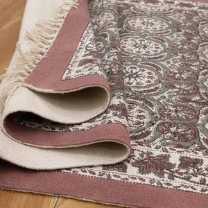 May include: Rolled-up area rug with a floral pattern in shades of gray, white, and burgundy. The rug has a cream-colored fringe and a burgundy border. The rug is made of a soft, woven material.
