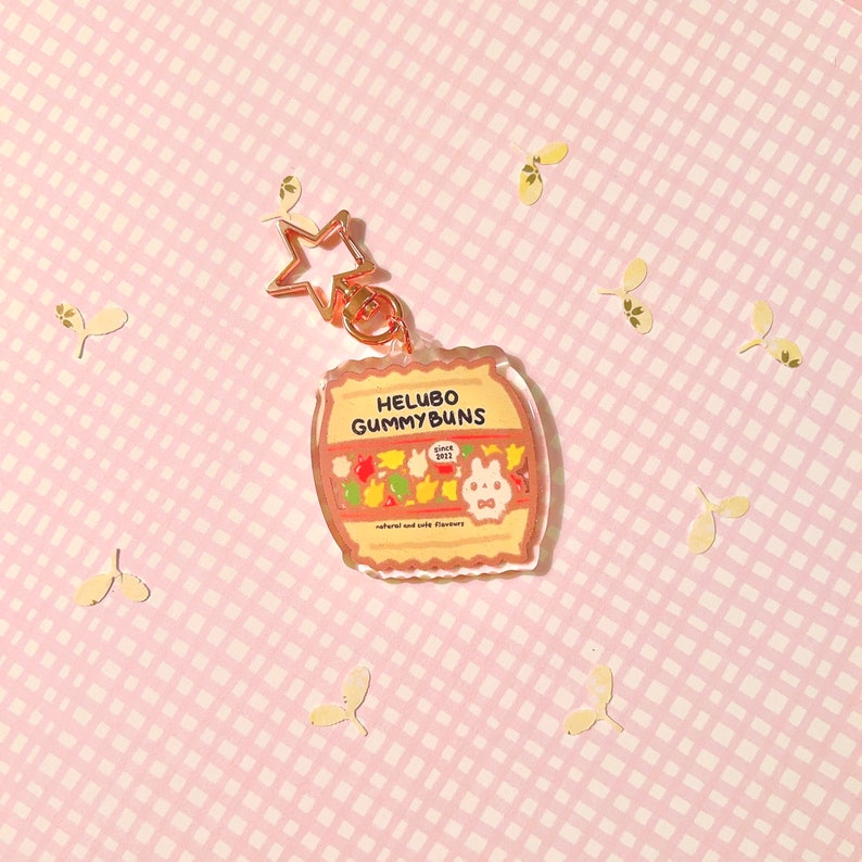 Pu&ograve; includere: Un portachiavi in ​​acrilico trasparente con un'illustrazione di un barile di caramelle gommose. Il barile &egrave; etichettato "Helubo Gummy Buns" con il testo "Since 2012" e "Natural and Wild Flavors". Il portachiavi ha un ciondolo a forma di stella dorata.