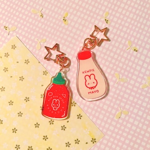 May include: Two clear acrylic keychain charms shaped like condiment bottles. One is red and says "Sriracha" with a cartoon bunny. The other is white and says "Kewpie Mayo" with a cartoon bunny. Both have gold star-shaped key rings.