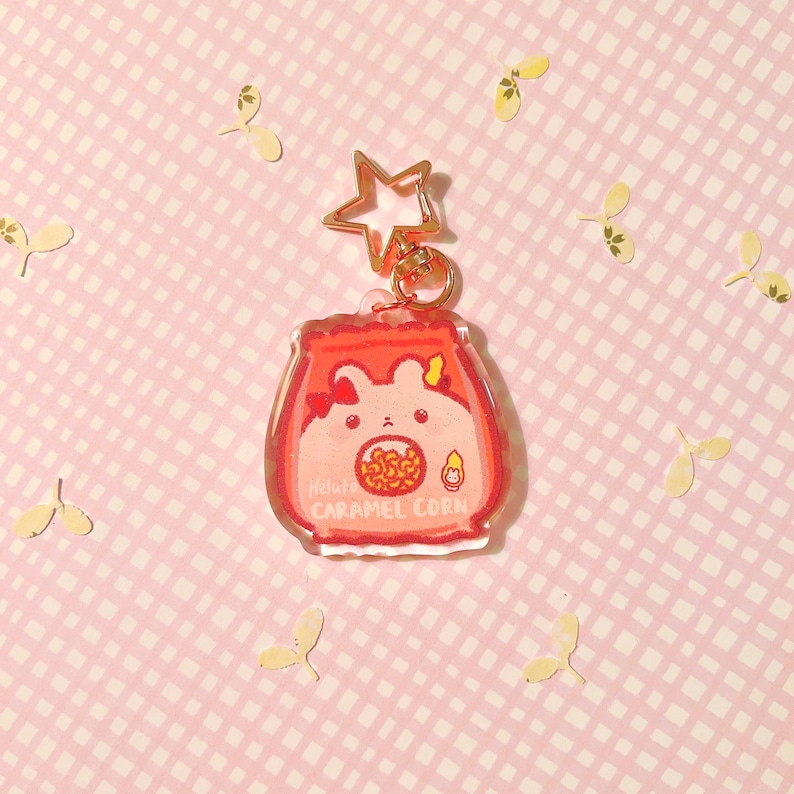 Pu&ograve; includere: Un portachiavi Hello Kitty rosa e bianco con un'immagine di Hello Kitty dei cartoni animati che tiene un sacchetto di popcorn al caramello. Il portachiavi &egrave; in acrilico trasparente e ha un ciondolo a forma di stella dorata.