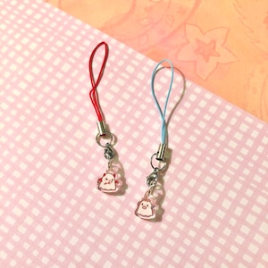 May include: Two small acrylic charms attached to thin loops. One charm features a red-horned ghost figure with a red cord, while the other displays a haloed ghost figure with a light blue cord. Both charms have silver-toned metal hardware.