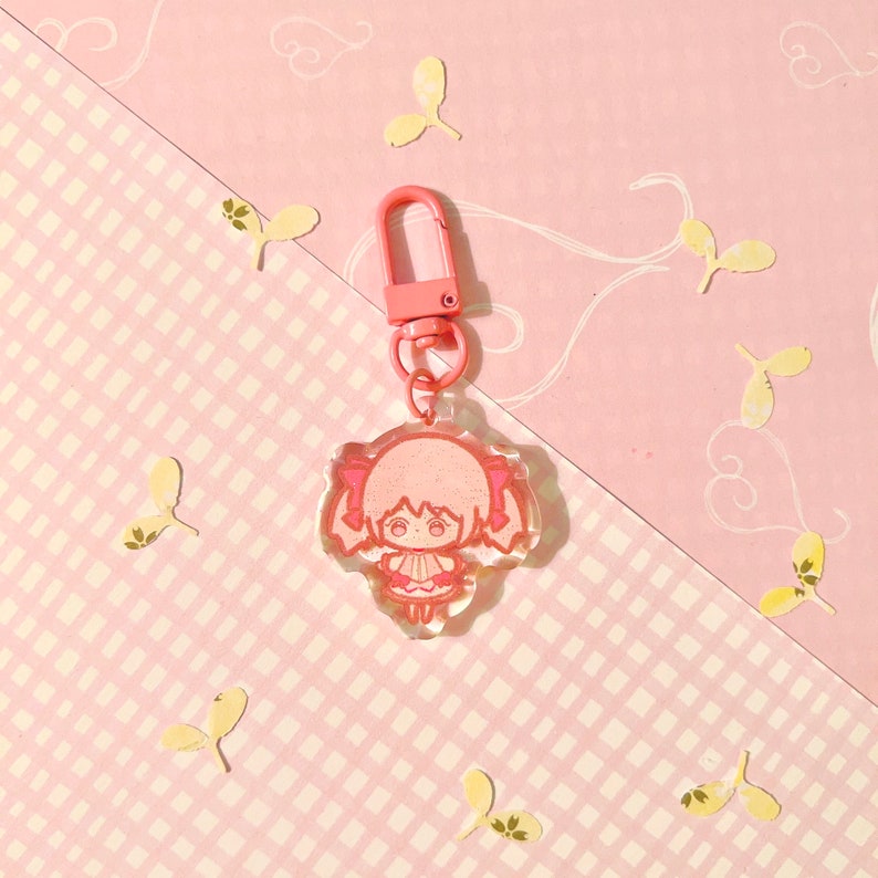 K&ouml;nnte beinhalten: Rosaer Anime-Charakter-Schl&uuml;sselanh&auml;nger mit Glitzerakzenten. Der Schl&uuml;sselanh&auml;nger zeigt ein kleines, cartoonartiges M&auml;dchen mit Z&ouml;pfen und einem rosa Kleid. Der Schl&uuml;sselanh&auml;nger ist an einem rosa Metallverschluss befestigt.