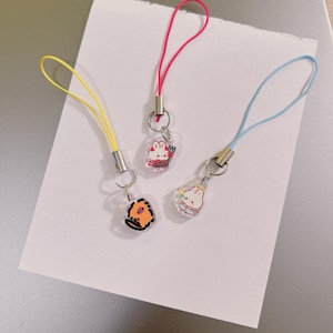 May include: Three acrylic phone charms featuring cartoon characters. One charm has a yellow cord and an orange creature, one has a pink cord and a white rabbit with a pitchfork, and one has a light blue cord and a white rabbit with a halo.