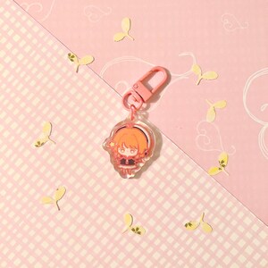 K&ouml;nnte beinhalten: Ein rosa und transparenter Acryl-Schl&uuml;sselanh&auml;nger mit einer Comicfigur mit orangefarbenen Haaren, die ein schwarz-wei&szlig;es Outfit tr&auml;gt. Die Figur h&auml;lt einen rosa Zauberstab mit einem gelben Stern am Ende.