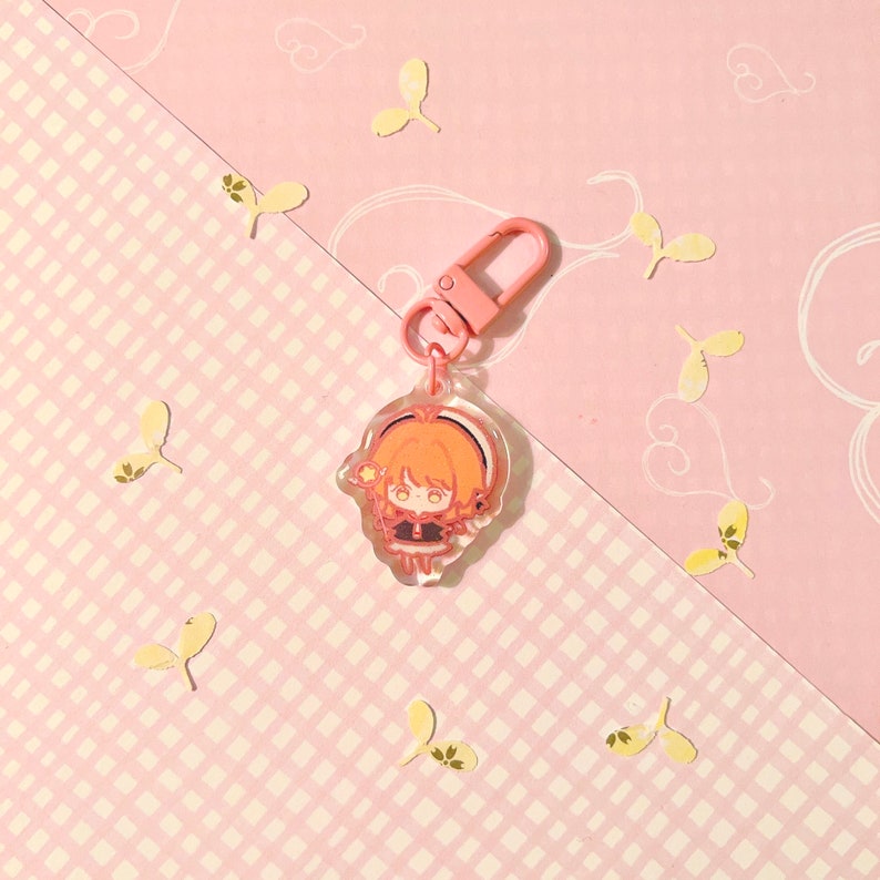 K&ouml;nnte beinhalten: Ein rosa und transparenter Acryl-Schl&uuml;sselanh&auml;nger mit einer Comicfigur mit orangefarbenen Haaren, die ein schwarz-wei&szlig;es Outfit tr&auml;gt. Die Figur h&auml;lt einen rosa Zauberstab mit einem gelben Stern am Ende.
