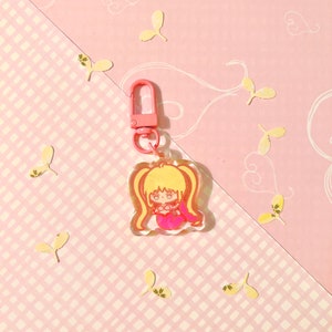 K&ouml;nnte beinhalten: Ein Schl&uuml;sselanh&auml;nger aus transparentem Acryl mit einer pinkfarbenen und gelben Comicfigur in Form einer Meerjungfrau. Der Schl&uuml;sselanh&auml;nger hat einen pinkfarbenen Metallverschluss.
