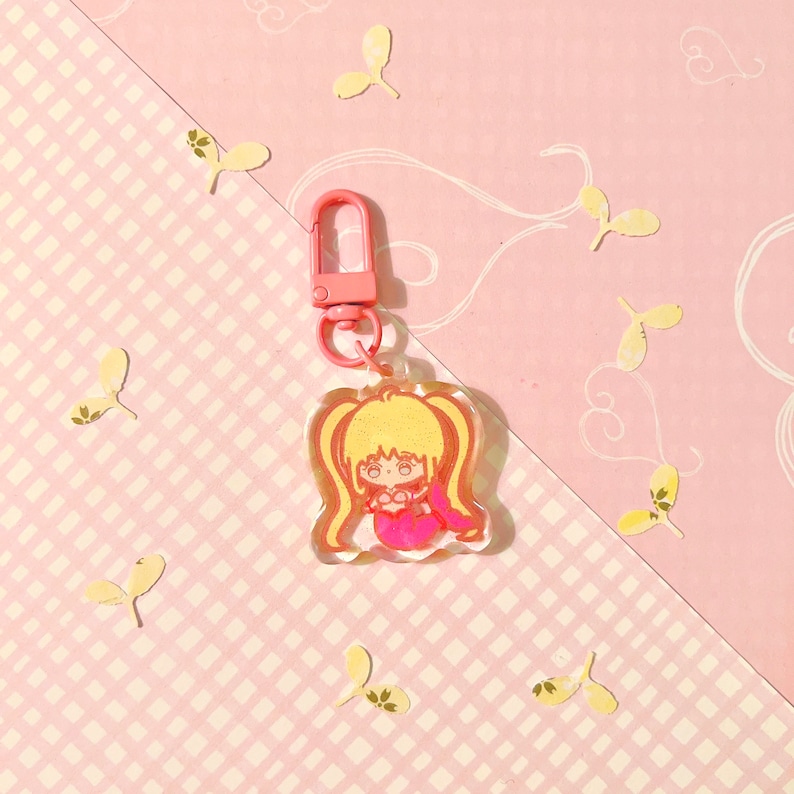 K&ouml;nnte beinhalten: Ein Schl&uuml;sselanh&auml;nger aus transparentem Acryl mit einer pinkfarbenen und gelben Comicfigur in Form einer Meerjungfrau. Der Schl&uuml;sselanh&auml;nger hat einen pinkfarbenen Metallverschluss.