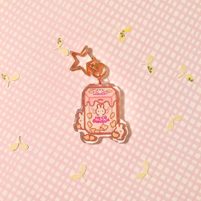 Pu&ograve; includere: Un portachiavi a forma di cartone di latte Hello Bunbun rosa e bianco con un ciondolo a stella dorata. Il portachiavi presenta un coniglio dei cartoni animati con naso e orecchie rosa, e il testo "Hello Bunbun" in lettere rosa.