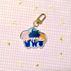 May include: A clear acrylic keychain with three anime-style characters in blue and red outfits. The keychain has a gold-coloured clasp and is set against a pink background with scattered yellow leaf-shaped confetti.