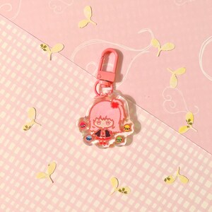 K&ouml;nnte beinhalten: Ein rosa und transparenter Schl&uuml;sselanh&auml;nger mit einem Cartoon-M&auml;dchen mit rosa Haaren und einem schwarzen Kleid. Der Schl&uuml;sselanh&auml;nger hat einen silbernen Metallclip und ist mit buntem Glitzer verziert.
