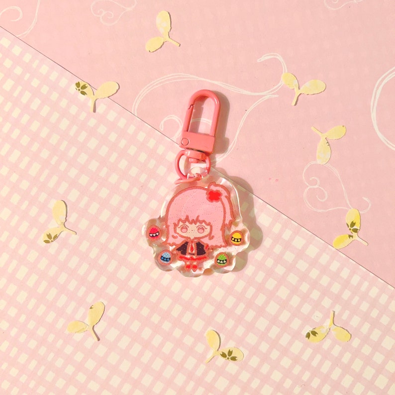 K&ouml;nnte beinhalten: Ein rosa und transparenter Schl&uuml;sselanh&auml;nger mit einem Cartoon-M&auml;dchen mit rosa Haaren und einem schwarzen Kleid. Der Schl&uuml;sselanh&auml;nger hat einen silbernen Metallclip und ist mit buntem Glitzer verziert.