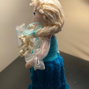 May include: A crocheted doll with long blonde hair, wearing a blue and white dress with a white crown on her head.