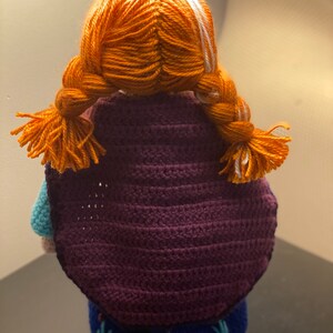 May include: A crocheted doll with orange hair in braids, wearing a purple cape and a blue dress with green embroidery.