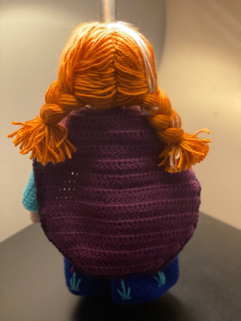 May include: A crocheted doll with orange hair in braids, wearing a purple cape and a blue dress with green embroidery.
