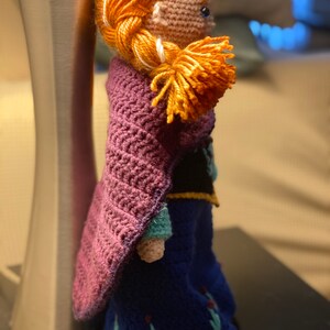 May include: A crocheted doll with long red hair, wearing a purple cape and a blue dress with floral embroidery. The doll is standing next to a silver lamp.