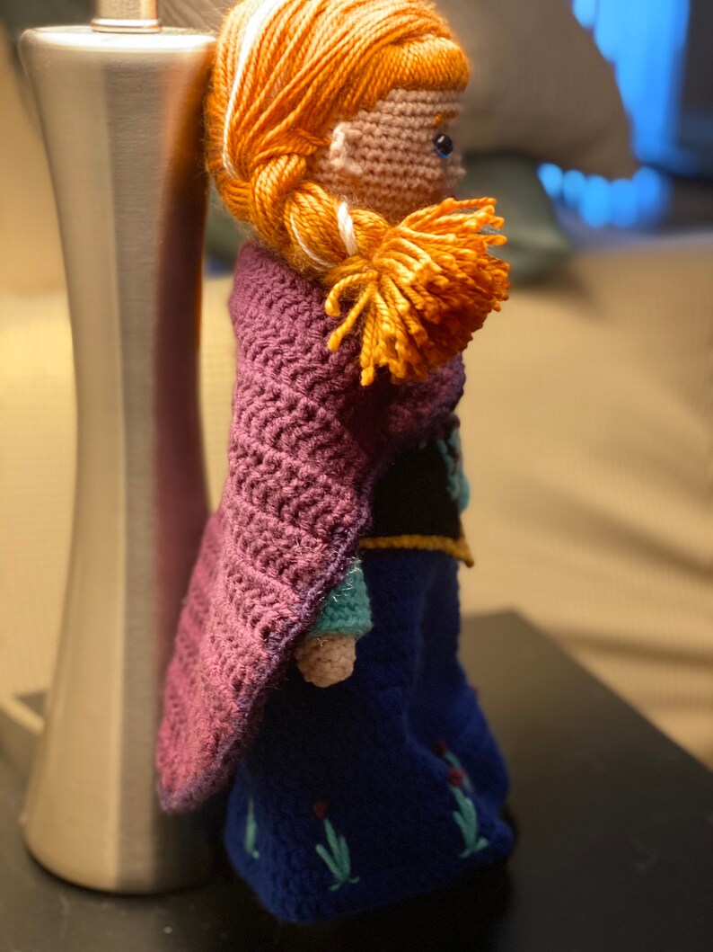 May include: A crocheted doll with long red hair, wearing a purple cape and a blue dress with floral embroidery. The doll is standing next to a silver lamp.