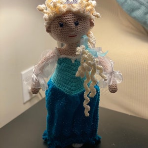 May include: A crocheted doll with blonde hair, a blue dress, and a silver crown. The doll is wearing a white tulle overlay on her dress and has a white crocheted braid.