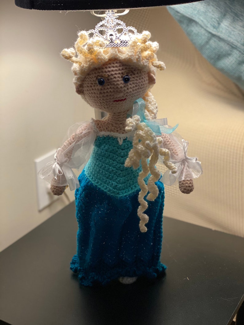 May include: A crocheted doll with blonde hair, a blue dress, and a silver crown. The doll is wearing a white tulle overlay on her dress and has a white crocheted braid.