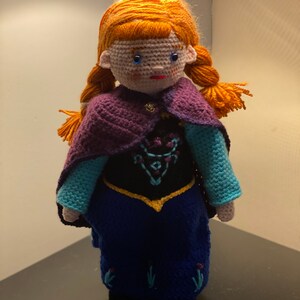 May include: A crocheted doll with red hair, blue eyes, and a purple cape. The doll is wearing a blue dress with a black bodice and gold trim.