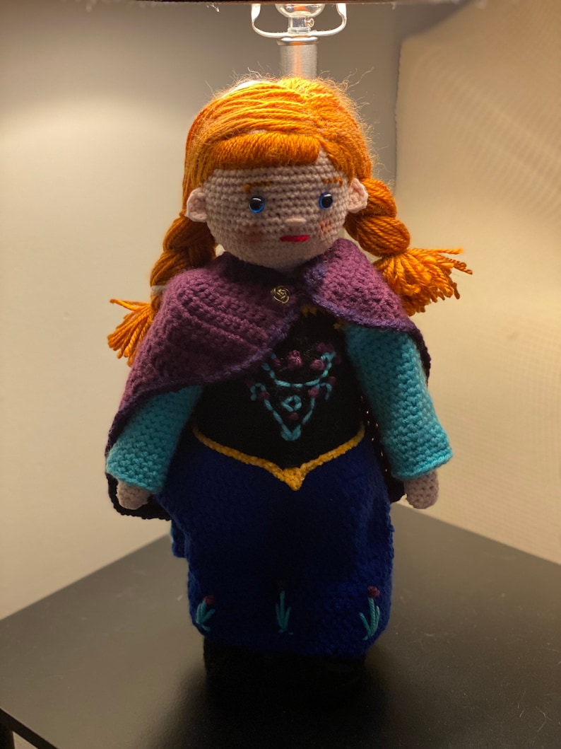 May include: A crocheted doll with red hair, blue eyes, and a purple cape. The doll is wearing a blue dress with a black bodice and gold trim.