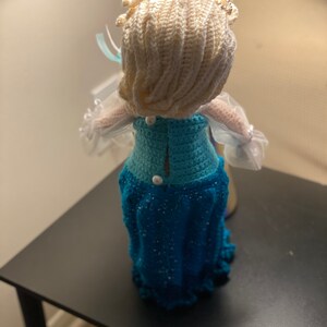 May include: A crocheted doll dressed in a blue and white dress with a white crocheted crown. The doll is standing on a black surface.