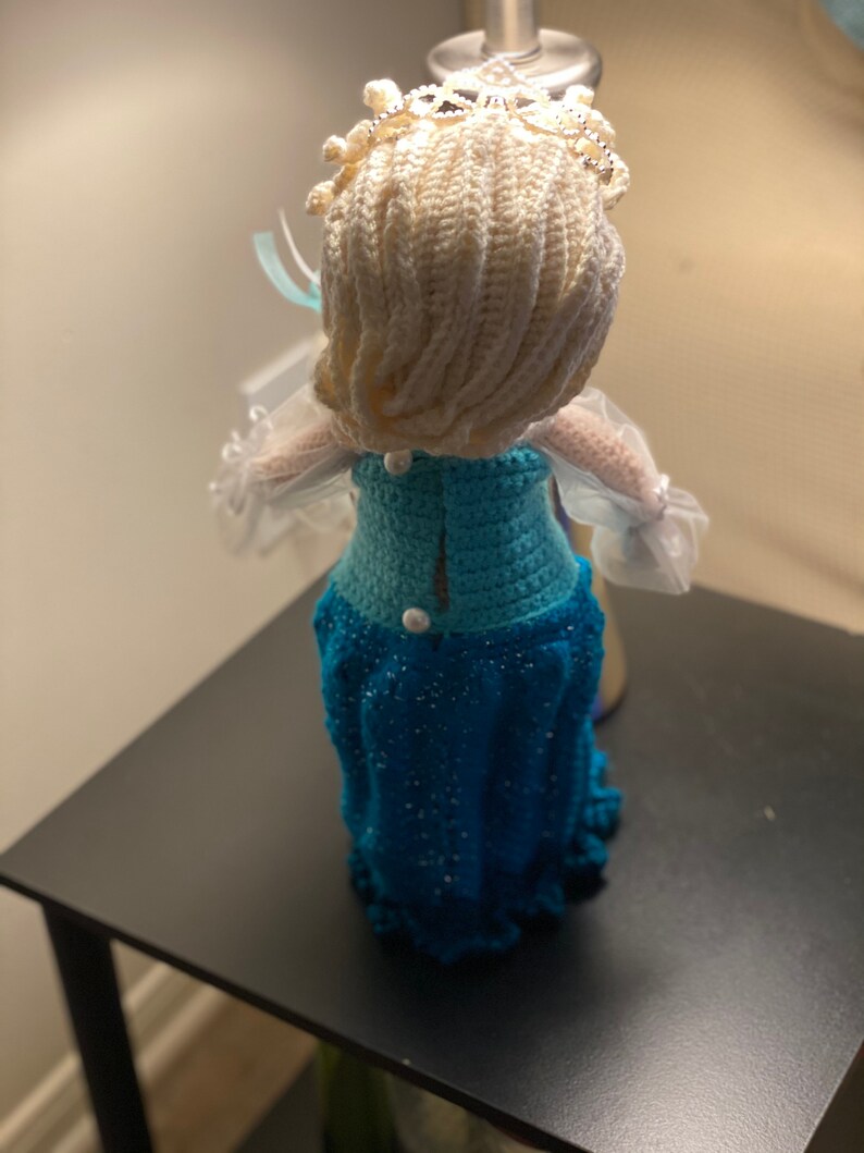 May include: A crocheted doll dressed in a blue and white dress with a white crocheted crown. The doll is standing on a black surface.