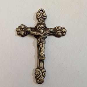 May include: A silver-toned metal crucifix pendant featuring a central figure of Jesus. The cross has ornate, rounded ends with embossed circular details and a small loop at the top for hanging. The metal has a weathered, textured finish throughout.