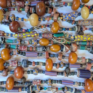 May include: A collection of multi-strand necklaces featuring a variety of antique trade beads. The strands include smooth amber-colored oval beads, patterned millefiori glass beads, striped cylindrical glass beads, and small textured spacers.