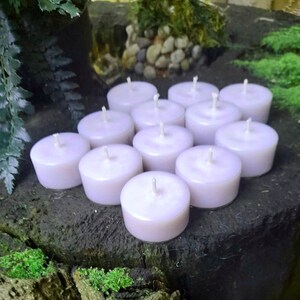 May include: A group of 12 white tea light candles arranged in a circle on a dark brown wooden surface. The candles are unscented and have a smooth, polished finish.