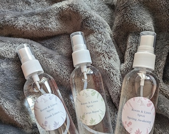 May include: Three clear glass spray bottles with white tops and labels. The labels are round and feature floral designs. The labels read "Room & Linen Spray" with different scents: "Fresh Linen", "Citrus Burst", and "Spring Awakening".