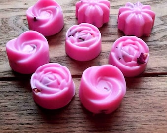 May include: Nine pink wax melts in various flower shapes, including roses and a star. The melts are on a wooden surface.