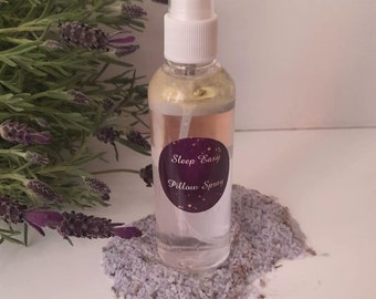 May include: A clear glass bottle with a white spray top and a purple label that says "Sleep Easy Pillow Spray". The bottle is sitting on a bed of lavender.